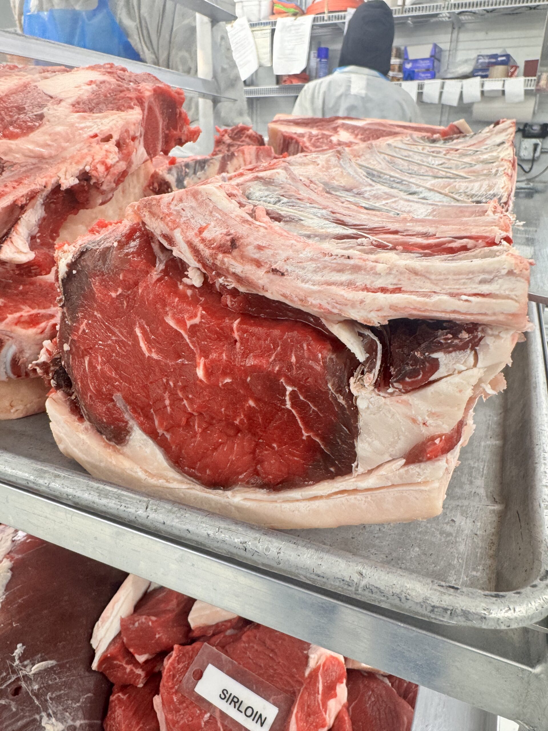 Fresh cuts of beef, including sirloin, displayed on a metal tray in a butcher shop. The image captures the marbled texture of the meat and the organized layout of various cuts in a clean, professional environment.